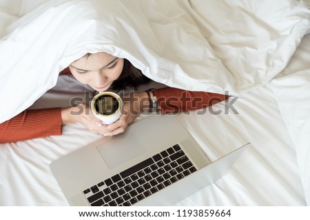 Charming young woman covering head with white blanket lying on bed and watching tv or movies streaming on laptop and holding a cub of hot coffee in winter season. Royalty-Free Stock Photo #1193859664
