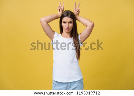 Fun and People Concept - Headshot Portrait of happy Caucasian woman with freckles smiling and showing rabbit ears with fingers over head . Pastel yellow studio background. Copy Space.