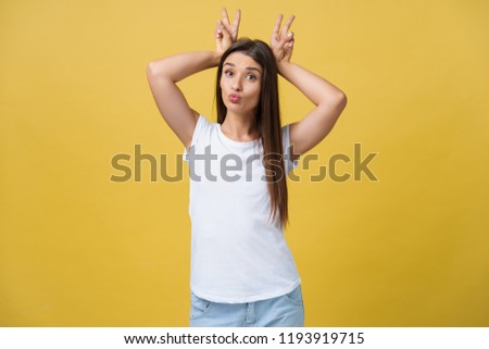 Fun and People Concept - Headshot Portrait of happy Caucasian woman with freckles smiling and showing rabbit ears with fingers over head . Pastel yellow studio background. Copy Space.