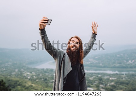 Young cheerful bearded guy makes selfie.Against the background of a young man a beautiful autumn landscape.Cheerful guy holds hands up.