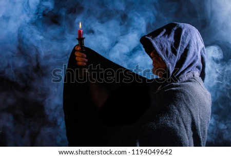 A man with a cloak and hood holds up a candleholder in the dark Royalty-Free Stock Photo #1194049642