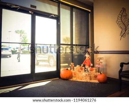 Beautiful Halloween decoration near office entrance from parking lots. Glass windows wall with natural light on scarecrow, colorful leaves, harvested orange pumpkins, squashes, gourd on hays