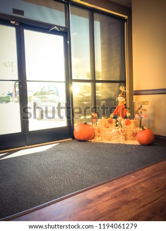 Vintage tone Halloween decoration near office entrance from parking lots. Glass windows wall with natural light on scarecrow, colorful leaves, harvested orange pumpkins, squashes, gourd on hays