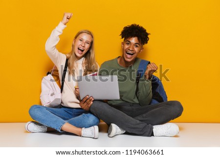 Photo of happy students man and woman 16-18 using silver laptop while sitting on floor with legs crossed isolated over yellow background