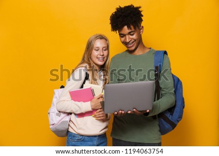 Photo of multiethnic students man and woman 16-18 wearing backpacks holding silver laptop and exercise books isolated over yellow background