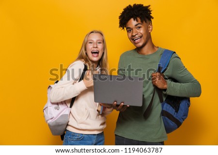Photo of smiling students guy and girl 16-18 wearing backpacks holding silver laptop and exercise books isolated over yellow background