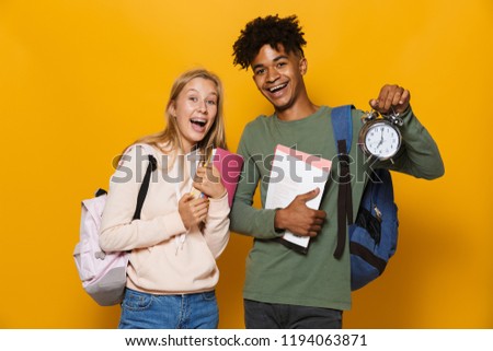 Photo of excited students man and woman 16-18 wearing backpacks holding exercise books and alarm clock isolated over yellow background