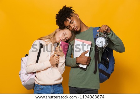 Photo of tired students man and woman 16-18 wearing backpacks sleeping while holding exercise books and alarm clock isolated over yellow background