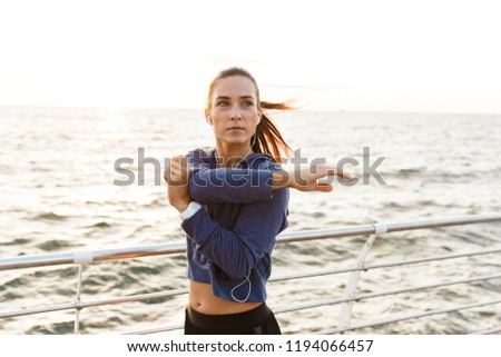 Photo of beautiful young sports woman outdoors on the beach listening music with earphones make stretching exercises.