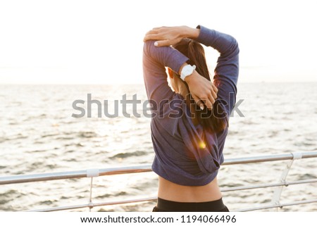 Back view picture of beautiful young sports woman outdoors on the beach listening music with earphones make stretching exercises.