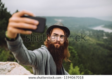 young bearded guy makes a selfie.Against the backdrop of a tourist green trees, mountains and river.Young guy has a stylish hat on his head and long hair.