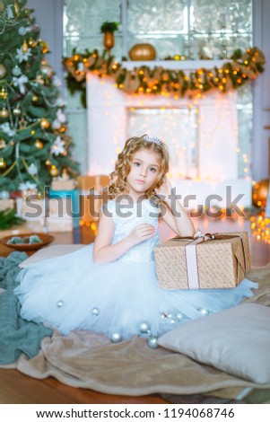 Cute little girl with curly blond hair at home near a Christmas tree with gifts and garlands and a decorated fireplace sitting on plaids and pillows
