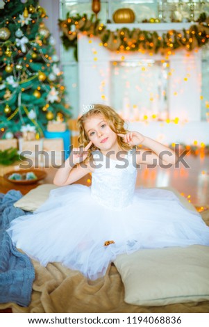 Cute little girl with curly blond hair at home near a Christmas tree with gifts and garlands and a decorated fireplace sitting on plaids and pillows
