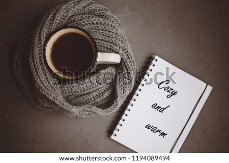 orange and yellow autumn leaves, a Cup of tea wrapped in a warm knitted scarf and Notepad with handle on gray background, top view, with copy space