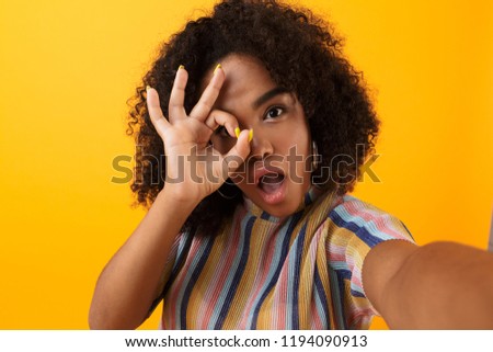 Image of beautiful young happy african cute girl posing isolated over yellow background take a selfie make okay gesture.
