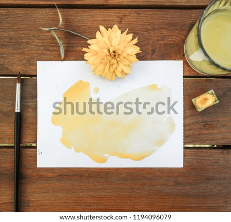 yellow watercolor on white paper, yellow flower, glass with yellow water, brush and yellow color on woody table