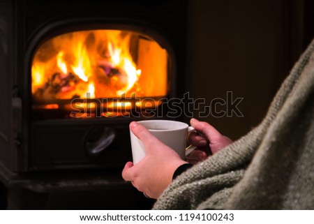 Woman having a hot cup of rink at the fireplace. Burning fireplace with wooden logs burning inside in the background. Warm light, romantic, christmas like atmosphere 