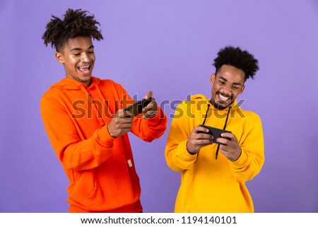 Two smiling african man friends dressed in colorful hoodies standing isolated over violet background, playing games on mobile phones