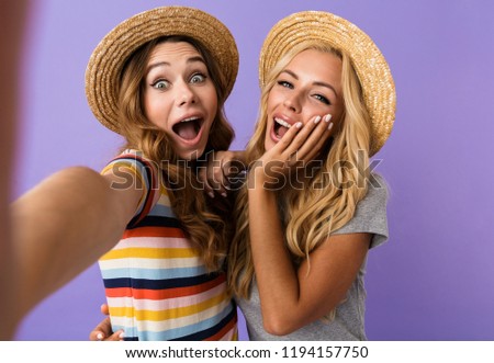 Two pretty excited young girls friends in summer hats standing isolated over violet background, taking a selfie with mobile phone