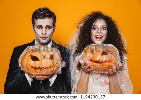 Happy bride and groom in wedding costumes with halloween make-up smiling to camera and holding pumpkin in web isolated over orange