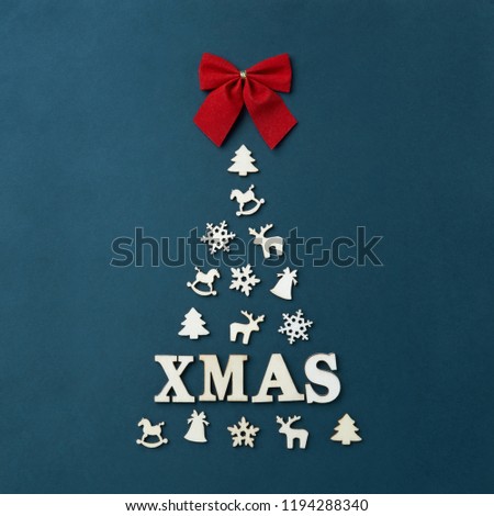 Flat lay top view Noel Christmas card with text XMAS. Large Christmas tree on dark blue background, lined with white wooden figures in the form of snowflakes, horse deer bell. Red bow. Winter holidays