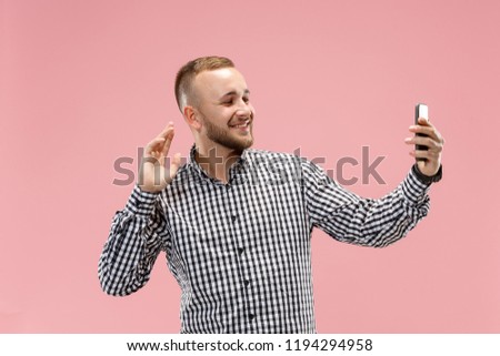 Portrait of attractive smiling young man taking a selfie with his smartphone. Isolated on pink studio background. Human emotions, facial expression concept. Trendy colors