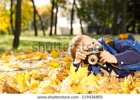 Small boy playing with a vintage slr camera lying down in colourful yellow autumn leaves outdoors in a park