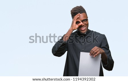 Young african american priest man over isolated background holding blank paper with happy face smiling doing ok sign with hand on eye looking through fingers