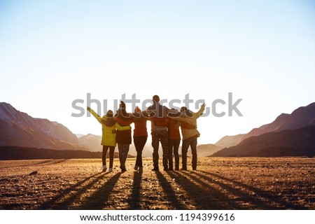 Group of happy friends stands and hugs against sunset mountains