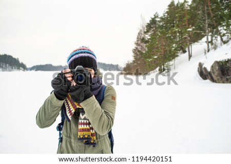 A woman photographer in warm clothes and a backpack is holding a camera in her hands and taking pictures of a winter landscape. Against the backdrop of lakes, cliffs and forests of the cold winter.