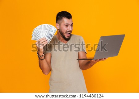 Portrait of a happy young casual man standing isolated over orange background, holding laptop computer, showing money banknotes
