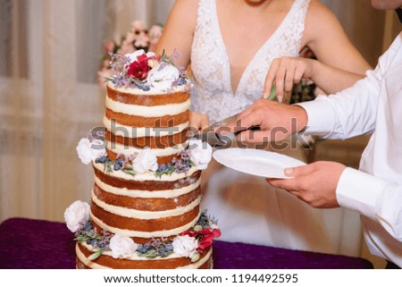 Close-up bride and groom hands cut multilevel white wedding cake decorated with flowers stands on a table. Concept of eating, sweets and desserts at a party.