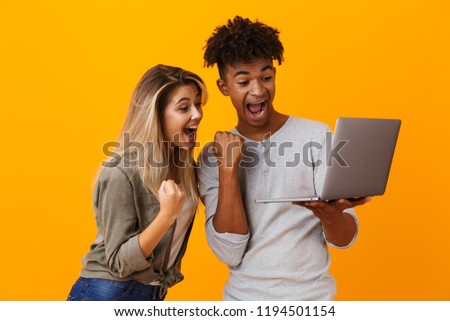 Photo of happy young loving couple standing isolated over yellow background using laptop computer make winner gesture.