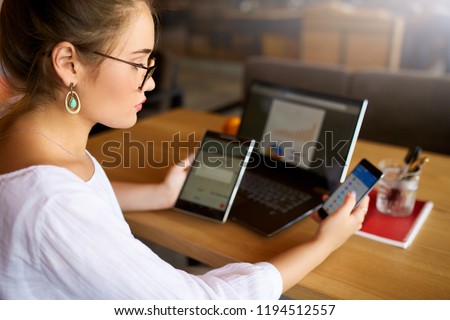Mixed race woman in glasses working with multiple electronic internet devices. Freelancer businesswoman has tablet and cellphone in hands and laptop on table with charts on screen. Multitasking theme Royalty-Free Stock Photo #1194512557