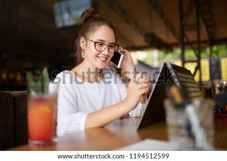 Mixed race freelancer working with convertible laptop and talking on cellphone with client in cafe. Asian caucasian businesswoman conducts negotiations via phone call. Multitasking business concept. Royalty-Free Stock Photo #1194512599