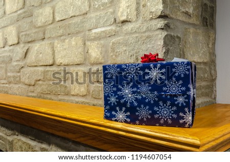 Chrismas present wrapped with blue wrapping paper with white snowflakes, with a red bow on top, sitting on a fireplace mantle