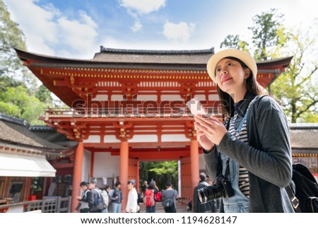 an elegant female traveler using her cell phone, standing in front of the temple and smiling happily