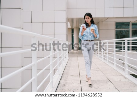 a full length photo of an elegant lady using phone while walking through the bridge, she is holding a cup of coffee