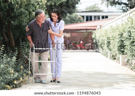 Nurse with patient using walker in retirement home. Young female nurse holding old man's shoulder in outdoor garden walking. Senior care, care taker and senior retirement home service concept. Royalty-Free Stock Photo #1194876043