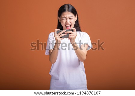 Woman playing mobile game isolated over orange background. Young asian woman in white t-shirt, upset mood. Young hipster lifestyle concept. Royalty-Free Stock Photo #1194878107