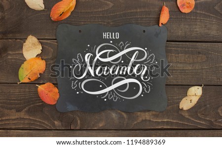 Chalkboard sign with hand lettering "Hello November". Autumn leaves and old wooden table on background.