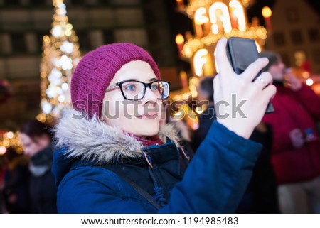 Woman in black glasses takes a photo, blurry lights on background. Christmas fair, outdoor. Holiday concert