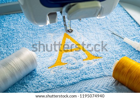 Close up picture of yellow alphabet logo A design and sewing accessories on blue towel in hoop of embroidery machine