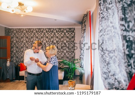 Mother is helping with a bow-tie to her son. Dressed and preparing her son for the wedding ceremony. The groom wears a jacket and looking to mother.