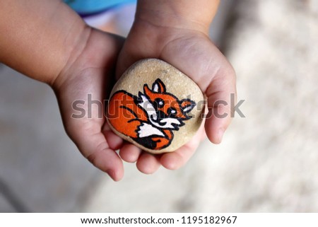 A little 3 year old toddler child's hands are gently holding a painted rock with a fox on it, that they will hide in the garden.