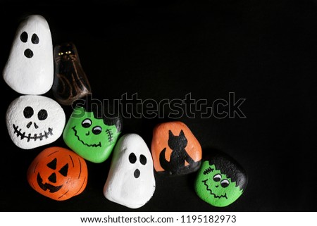 A collection of hand-painted spooky cartoon halloween painted craft rocks featuring ghosts, pumpkins, cats, skeleton, and monsters are bordering a black background with copy-space.