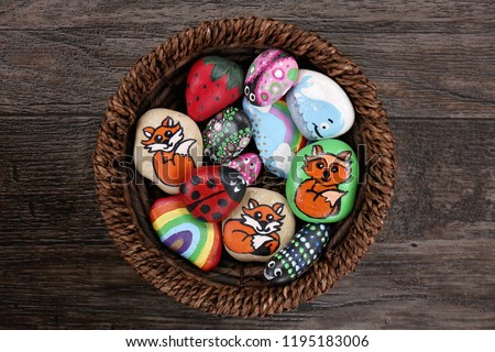 A collection of fun, handpainted, colorful cartoon rocks are together in a wicker basket, on a wooden plank background.