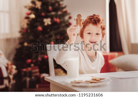 Sweet childhood. Portrait of joyful little child showing you a cookie while sitting at the table and having breakfast