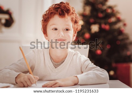 Positive mood. Close up of smiling little child sitting at the table while holding a pencil and looking away