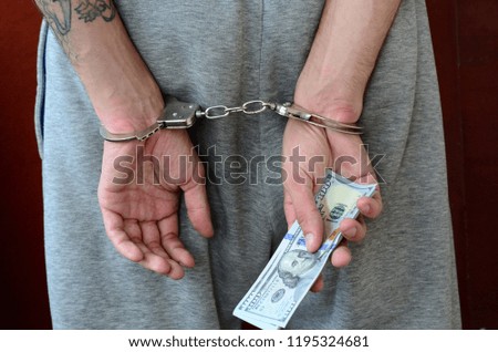 An arrested guy in gray pants with handcuffed hands holds a huge amount of dollar bills. Back view